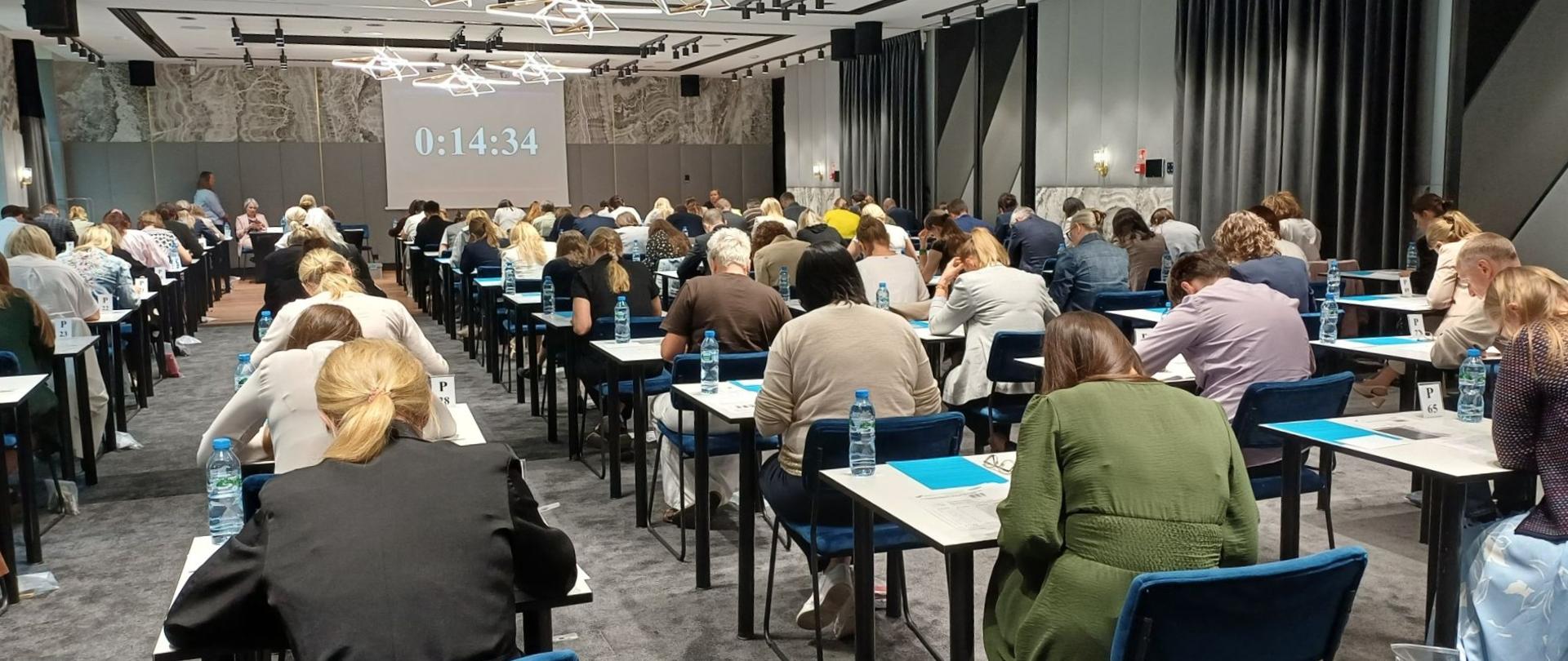 kilkadziesiąt osób pisze egzamin w dużej sali z wyświetlonym zegarem