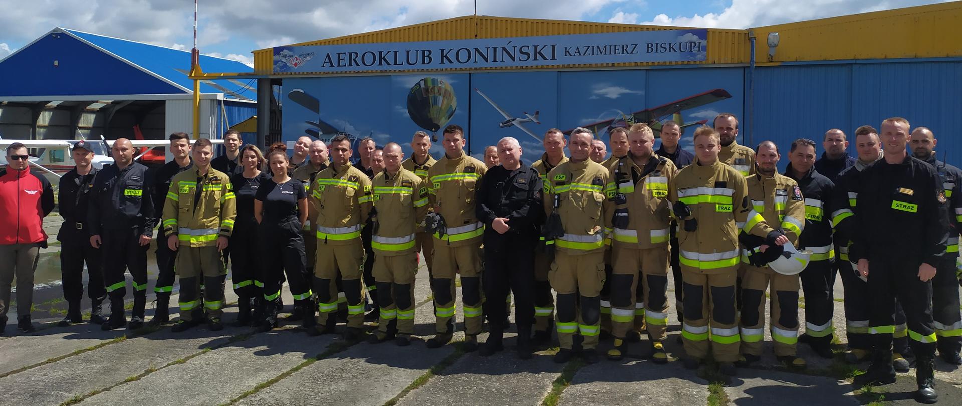 Grupa około 30 strażaków w mundurach stoi w w dwuszeregu przed hangarami lotniczymi. W tle widać samolot stojący przed hangarem oraz napis Aeroklub Koniński Kazimierz Biskupi