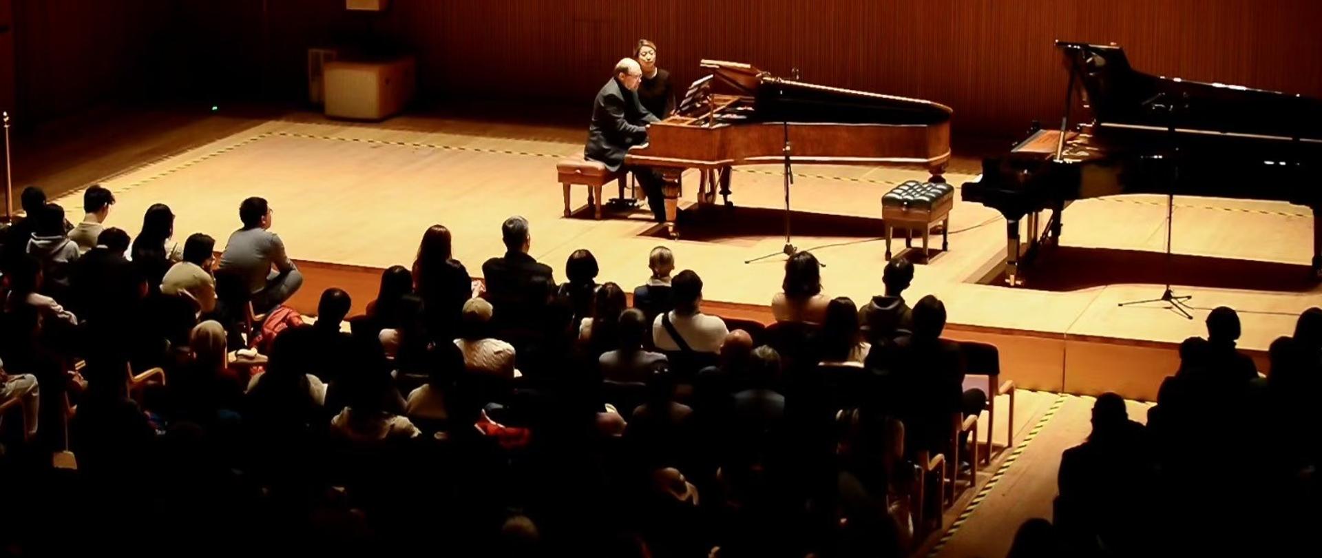 Mężczyzna i kobieta siedzą przy fortepianie na scenie, przed nimi tonąca w mroku widownia