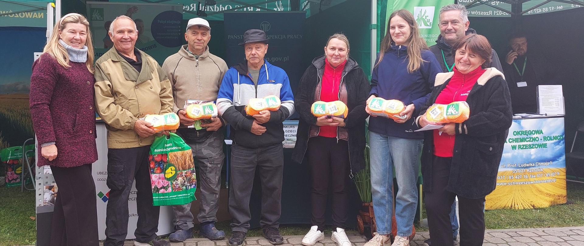 Przed stoiskiem prewencyjnym Kasy znajduje się grupa osób. Laureaci konkursu – trzy kobiety i trzech mężczyzn – trzymają w rękach nagrody opatrzone logo KRUS.