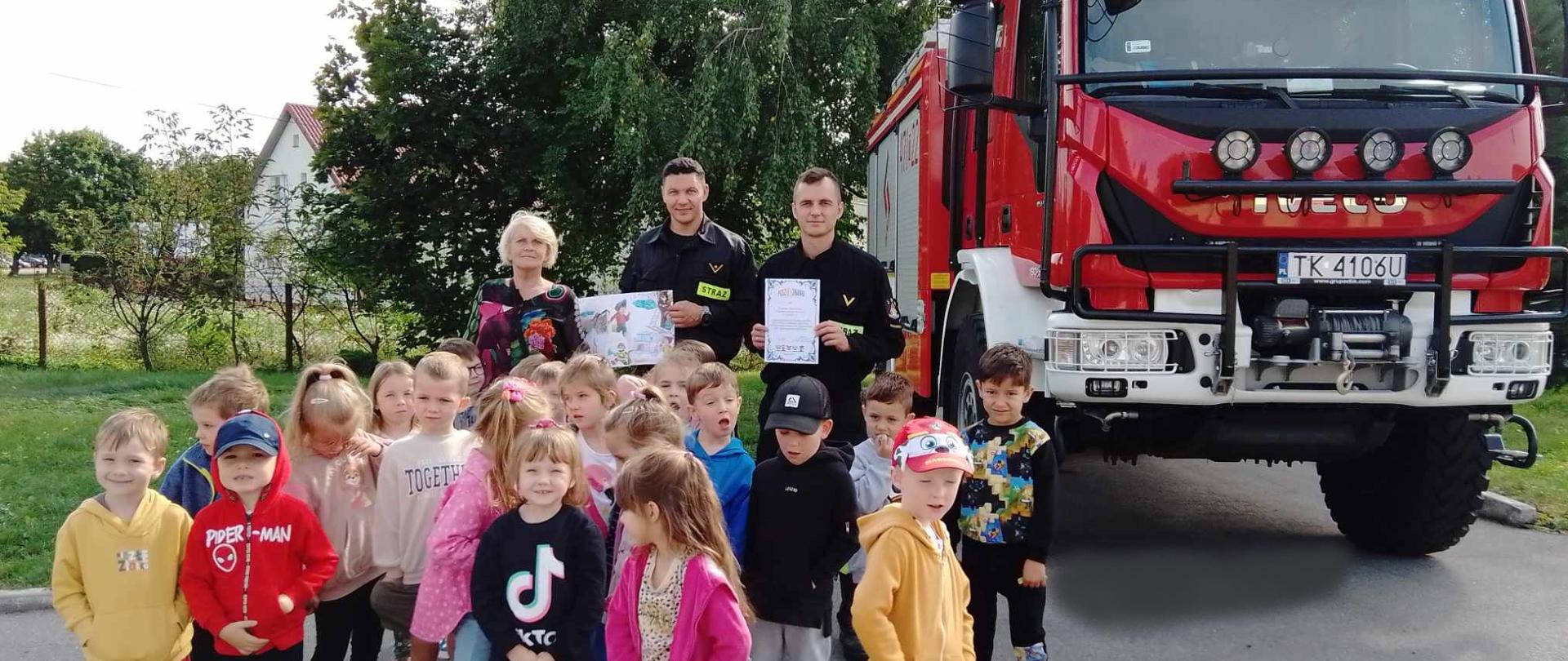 Zdjęcie przedstawia grupę dzieci z przedszkola wraz ze strażakami na tle samochodu strażackiego. 