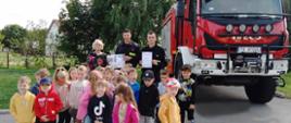 Zdjęcie przedstawia grupę dzieci z przedszkola wraz ze strażakami na tle samochodu strażackiego. 