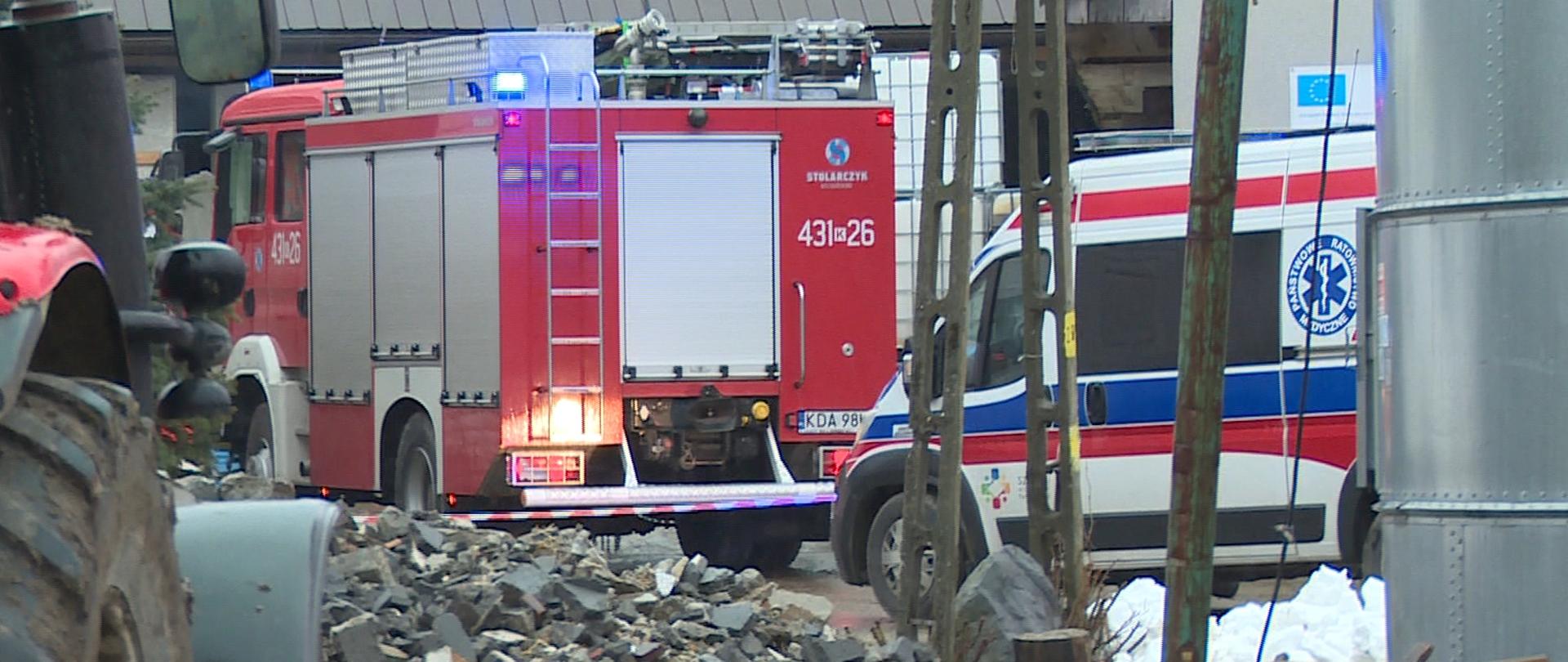 Na zdjęciu widać w dalszym planie samochód pożarniczy oraz Zespół ratownictwa medycznego, stojące przed budynkami, na zdjęciu widoczny jest także ciągnik oraz sterta gruzu.