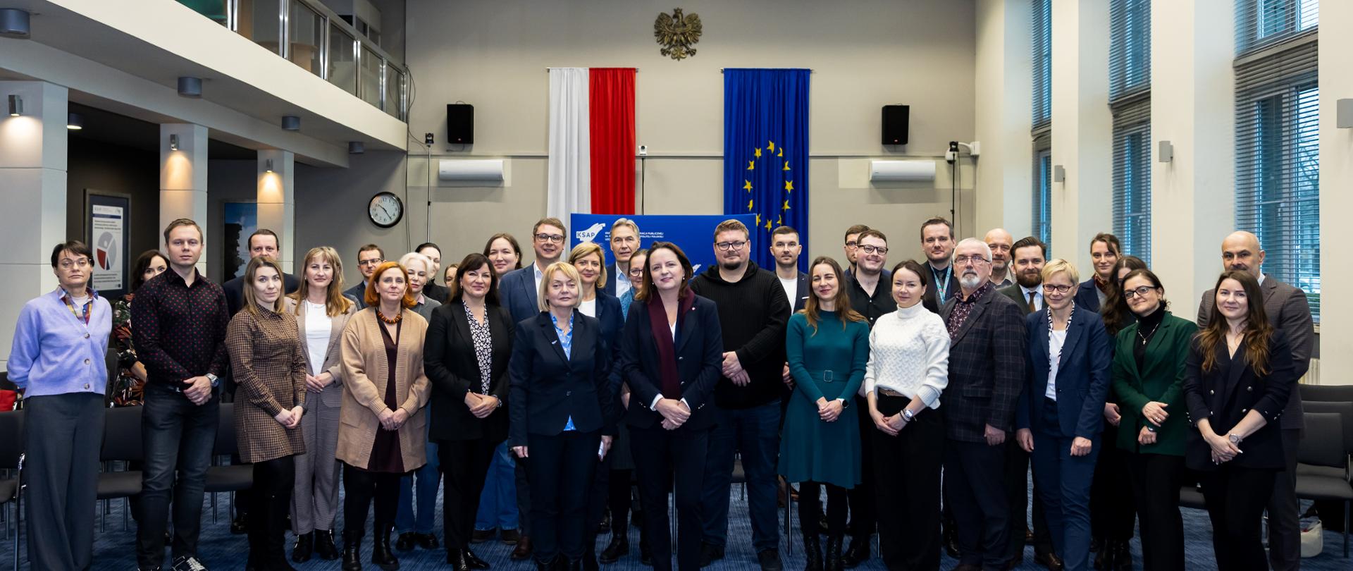 Grupa kilkunastu osób na tle flag: Polski i Unii Europejskiej. 