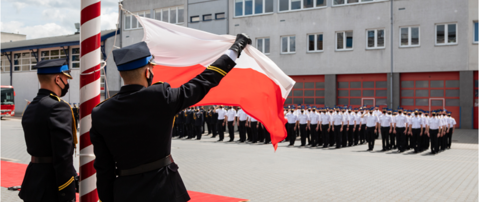 maszt z flagą Polski, obok dwóch strażaków, w tle budynek oraz poddodziały strażaków