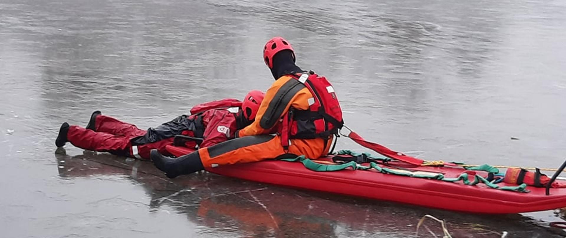Strażak w skafandrze wypornościowym podejmuje przy użyciu sań lodowych osobę poszkodowaną z przerębla na terenie akwenu zalodzonego. 