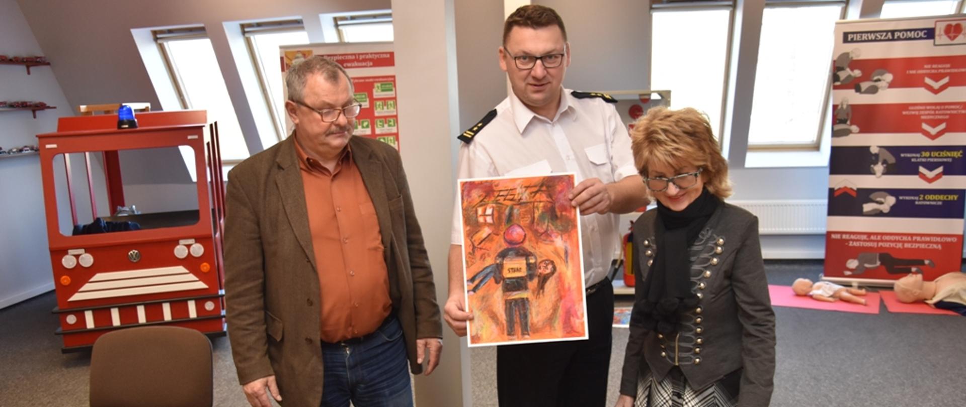 Komendant Powiatowy PSP w Ostródzie bryg. Tomasz Ostrowski trzyma zwycięską pracę Ogólnopolskiego Konkursu Plastycznego „Zapobiegam pożarom”.