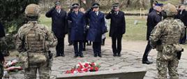 Uczestnicy ceremonii upamiętniającej oddają hołd przy grobie; wśród obecnych znajdują się przedstawiciele służb mundurowych w galowych mundurach, w tym Państwowej Straży Pożarnej oraz Policji, po obu stronach grobu stoją żołnierze w umundurowaniu bojowym zwróceni do osób oddających hołd