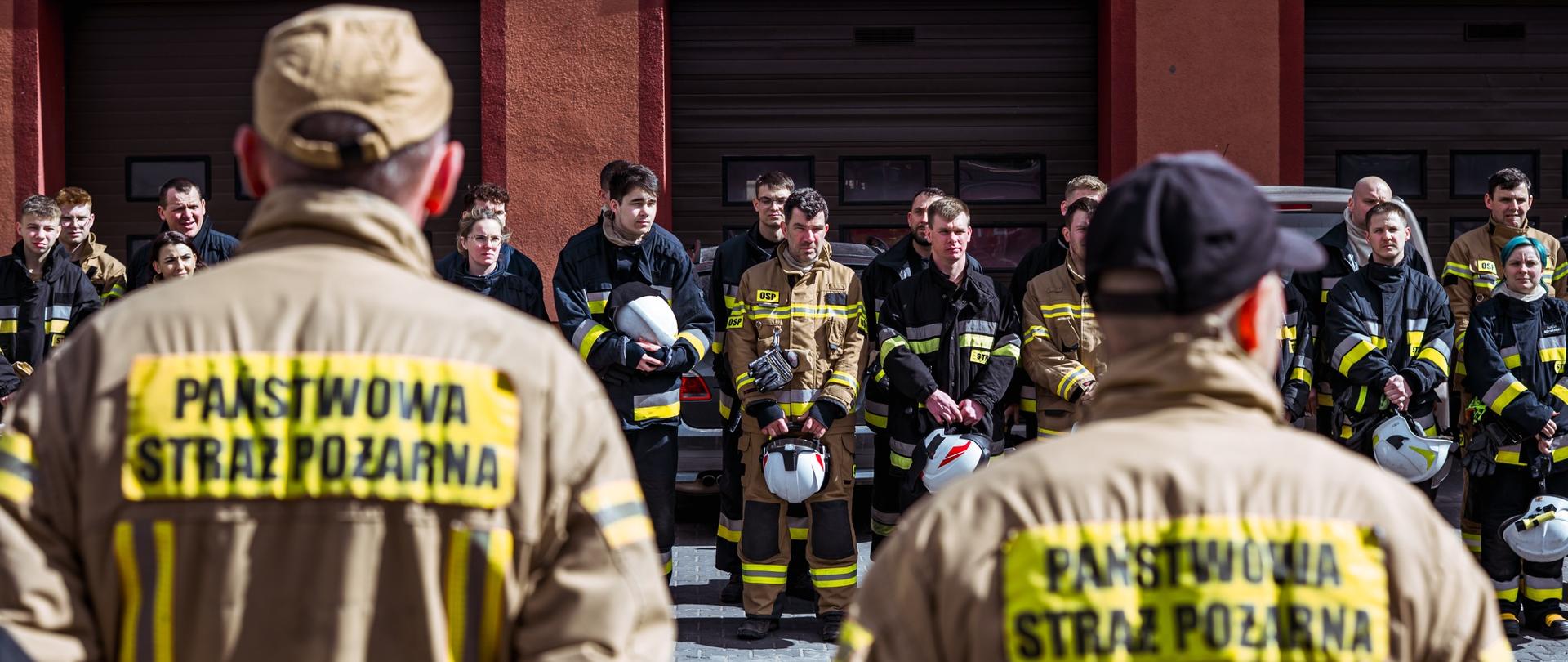 Grupa strażaków stoi ustawiona w szeregu przed budynkiem remizy z zamkniętymi bramami garażowymi. Na pierwszym planie widać dwóch strażaków tyłem do kamery, w kurtkach z napisem „Państwowa Straż Pożarna”. Pozostali strażacy trzymają hełmy w rękach i słuchają, jakby uczestniczyli w zbiórce lub odprawie.