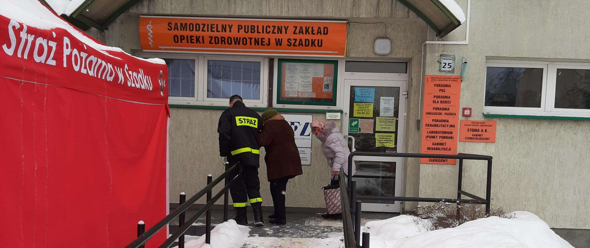 Na zdjęciu widoczny jest strażak OSP w ubraniu koszarowym, który pomaga osobie wejść do samodzielnego publicznego zakładu opieki zdrowotnej w Szadku gdzie prowadzona jest akcja szczepień przeciwko COVID-19. Po lewej stronie widoczny czerwony namiot z napisem OSP Szadek. 