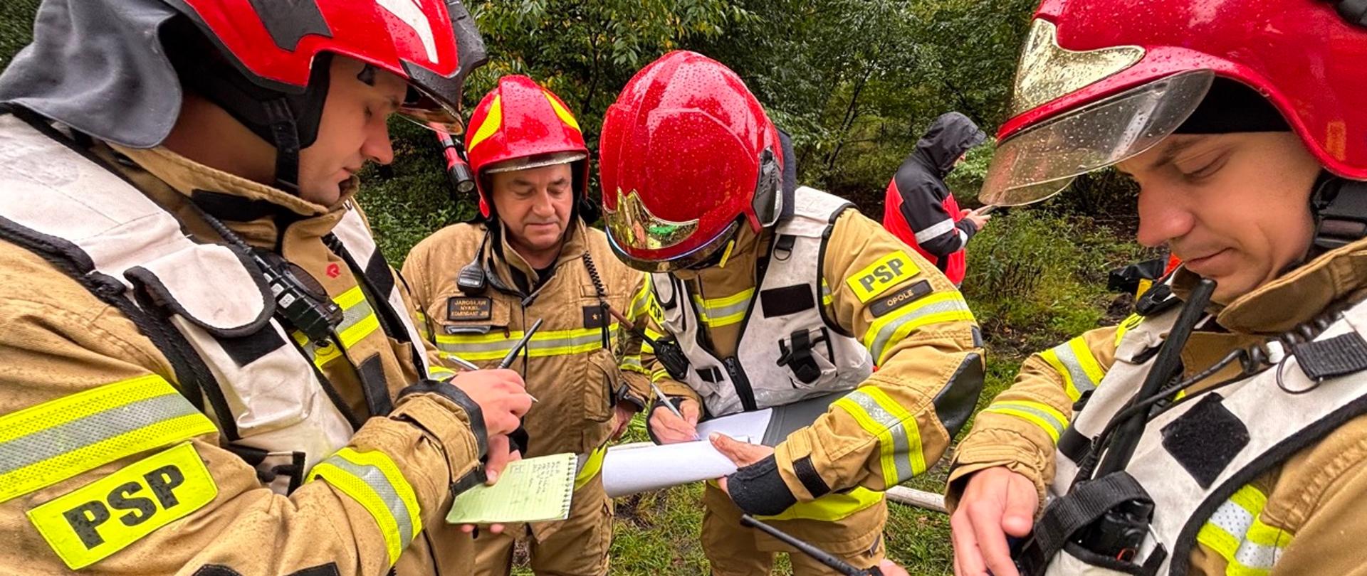 Grupa strażaków w ubraniach ochronnych i czerwonych hełmach uczestniczy w ćwiczeniach ratowniczych ICSO Chemical 2025 w dniu 6 października 2025 roku. Strażacy ustalają wspólnie strategię dalszych działań.
