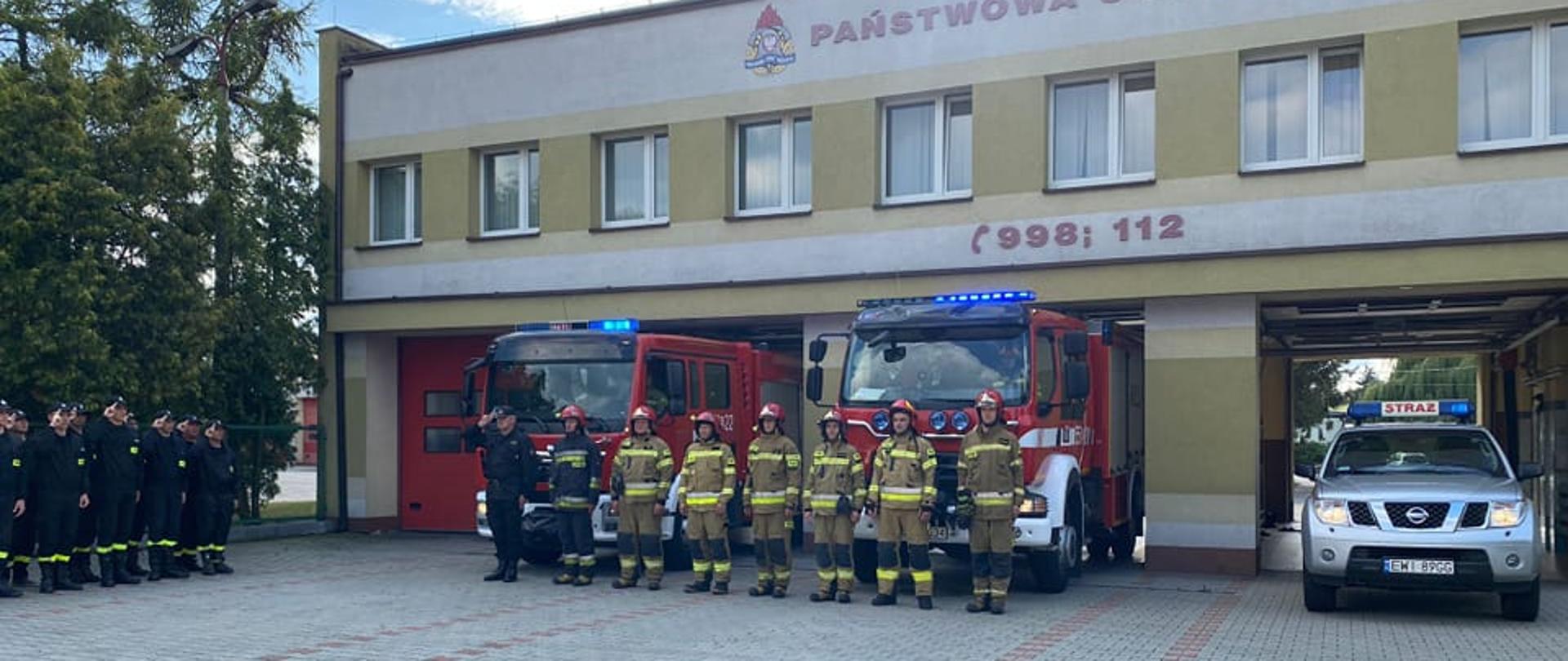 Zdjęcie przedstawia 8 strażaków państwowej straży pożarnej w ubraniach bojowych stojących na tle trzech samochodów pożarniczych i budynku komendy straży w wieluniu oraz stojących obok druhów osp