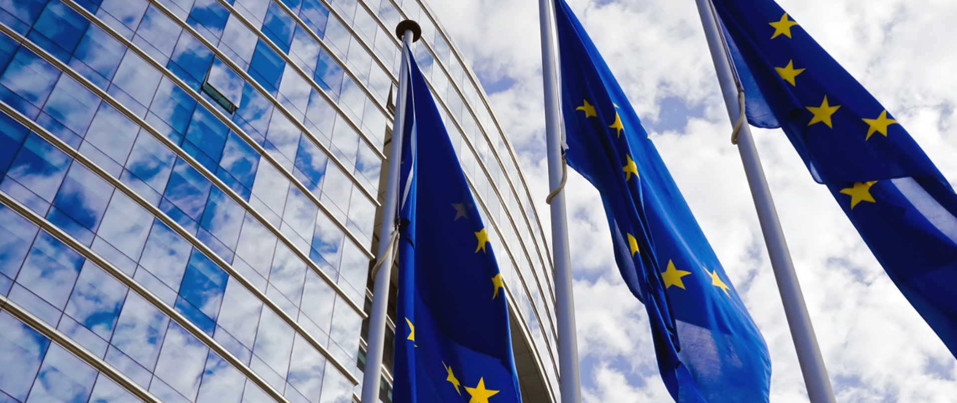 Zdjęcie przestawia budynek Rady Unii Europejskiej i 3 maszty z flagami EU 