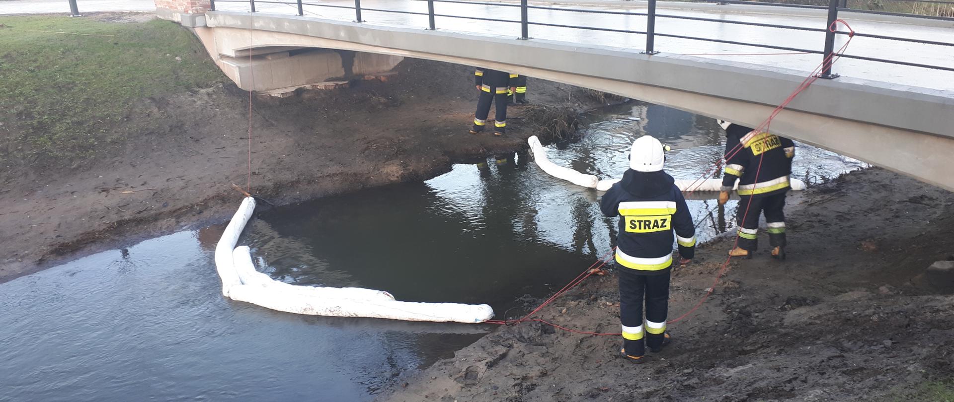 Na zdjęciu widać czterech strażaków, ubranych w stroje strażackie, znajdujący się pod małym mostkiem, pod którym płynie rzeczka ( po dwóch strażaków na jeden brzeg rzeki). Na rzece widać rozłożone 2 rękawy sorpcyjne .