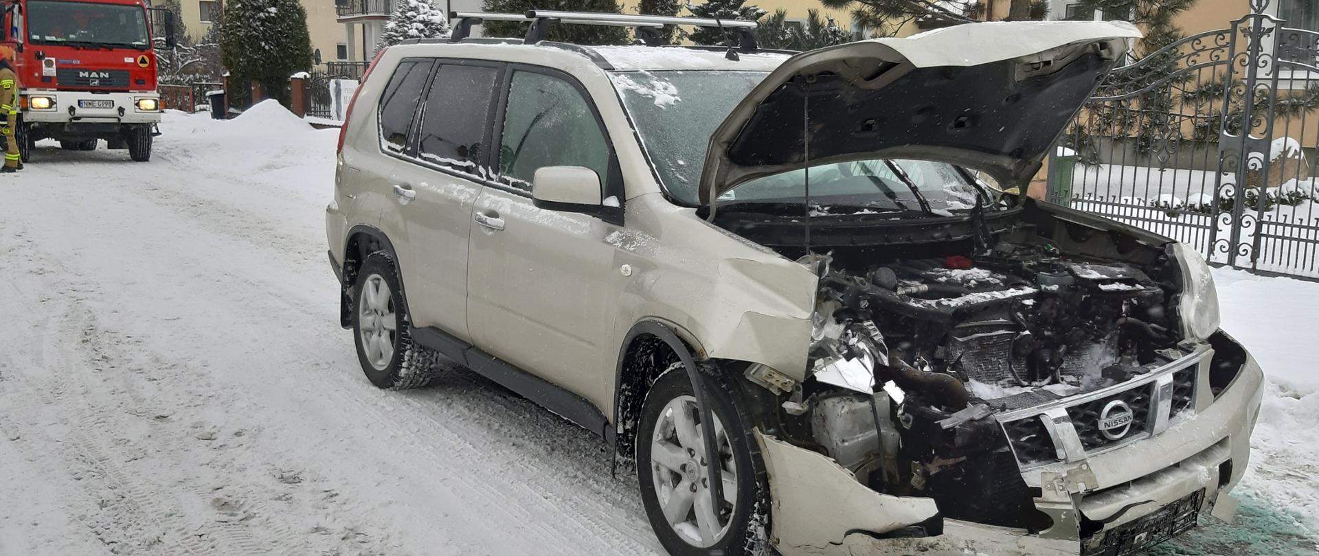 Rozbity przód samochodu marki Nissan X Trial z otwartą maską bagażnika, za pojazdem w oddali widać strażaka, który stoi przy samochodzie gaśniczym , a także za pojazdami znajdują się domy jednorodzinne. 