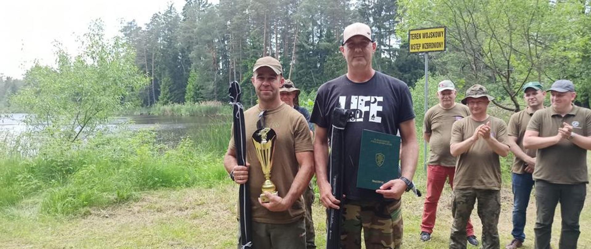 Na pierwszym planie dwóch mężczyzn w czapeczkach z daszkiem trzymają w jednej ręce wędki w pokrowcach a drugiej nagrody. Z tyłu kilka osób bija brawo.