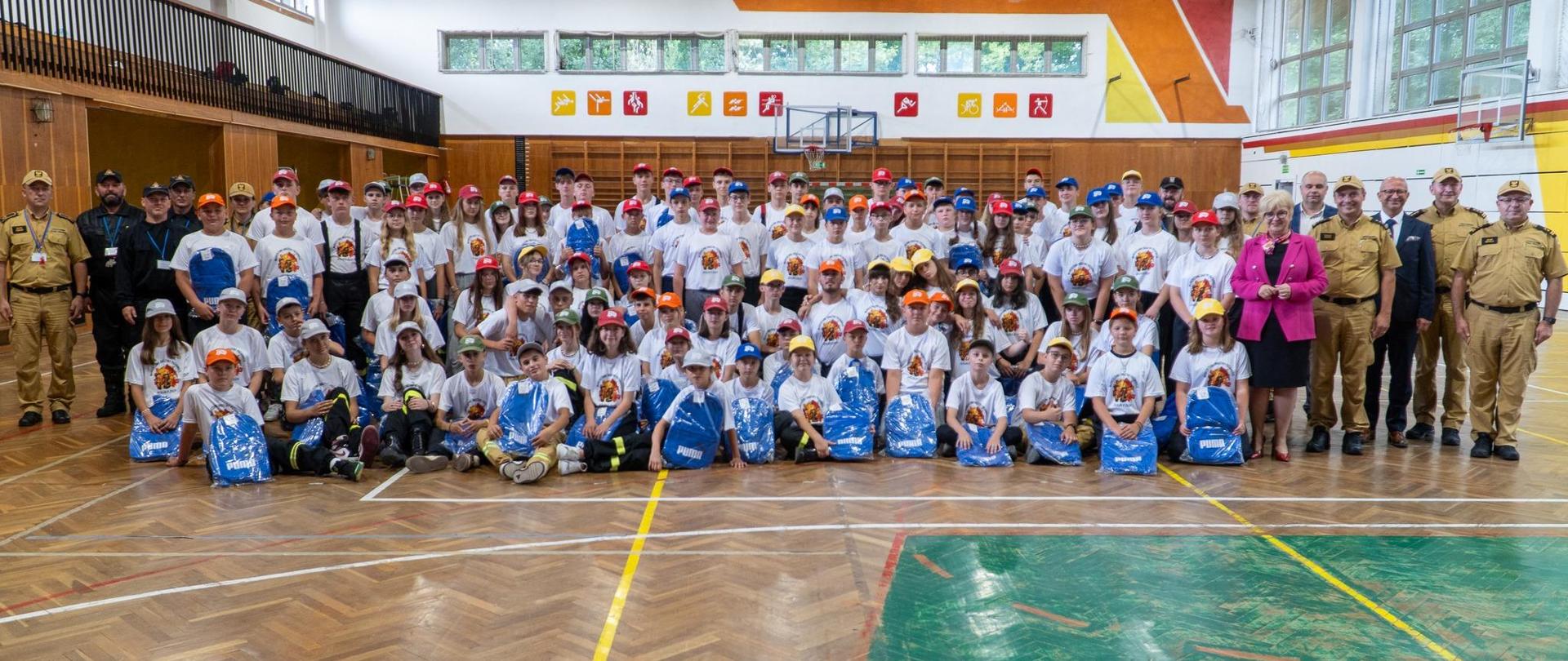 Zdjęcie zrobione na w sali gimnastycznej, podczas uroczystej zbiórki uczestników obozu Młodzieżowych Drużyn Pożarniczych. Zdjęcie grupowe uczestników obozu oraz funkcjonariuszy Państwowej Straży Pożarnej. 