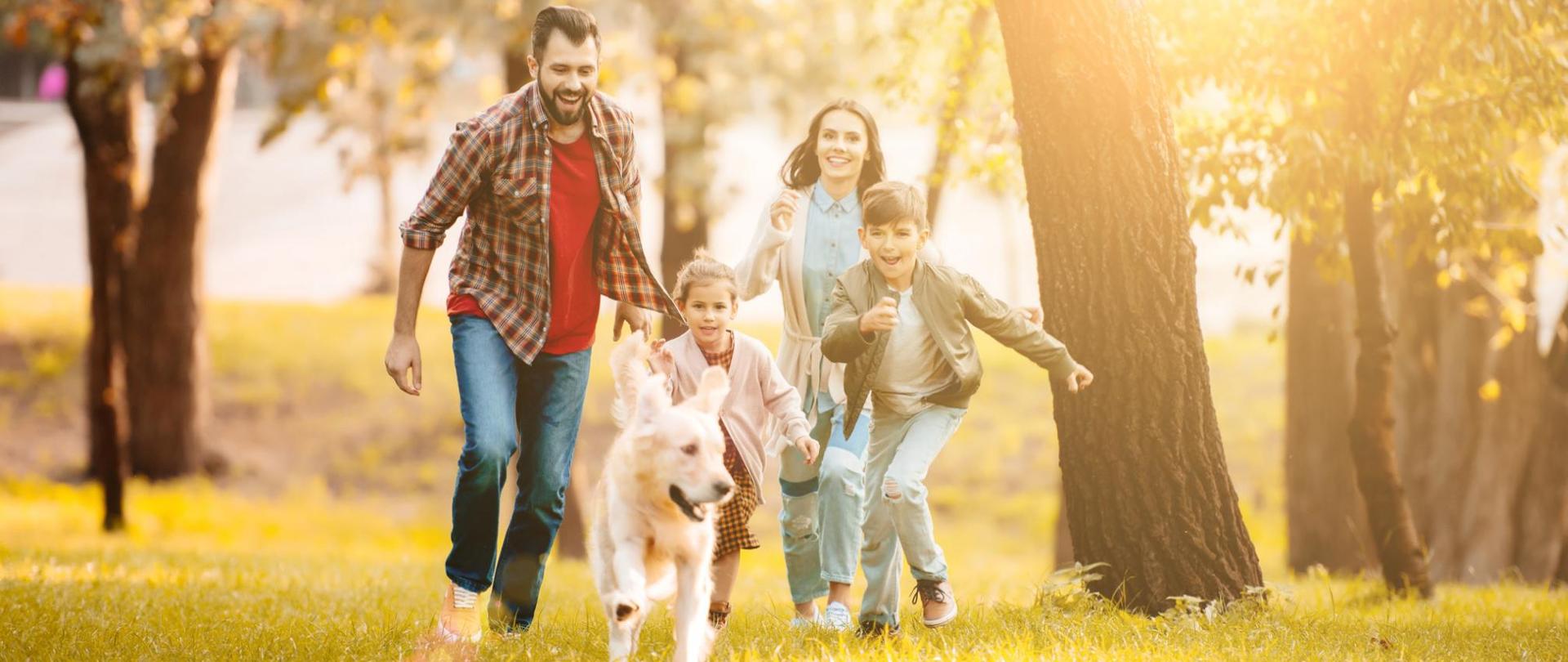 Na zdjęciu jest biegnąca rodzina wraz z psem w parku.
