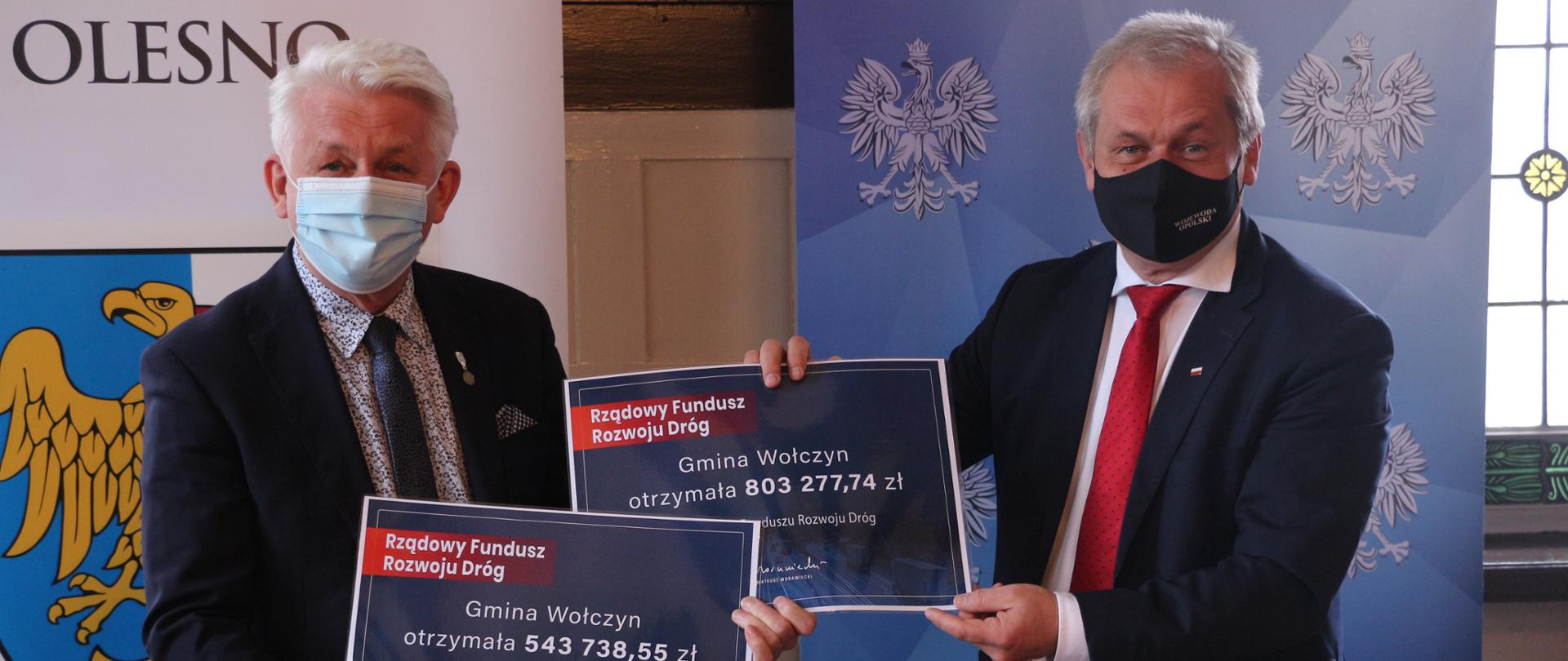 Na zdjęciu dwóch mężczyzn, stojących koło siebie. Od lewej burmistrz Wołczyna i wojewoda opolski. Mężczyźni pozują do zdjęcia, trzymają w dłoniach czeki (każdy po jednym) na realizację inwestycji drogowych. Czeki to drugi w formacie 30 na 40 cm, koloru granatowego z wpisaną kwotą dofinansowania. W tle dwa roll upy: po prawej wojewody, po lewej gminy Olesno. 
