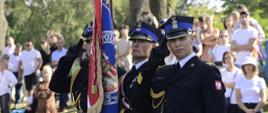 Pod zielonymi drzewami stoi strażacki poczet sztandarowy - troje strażaków w czarnych galowych mundurach, za nimi grupa ludzi.