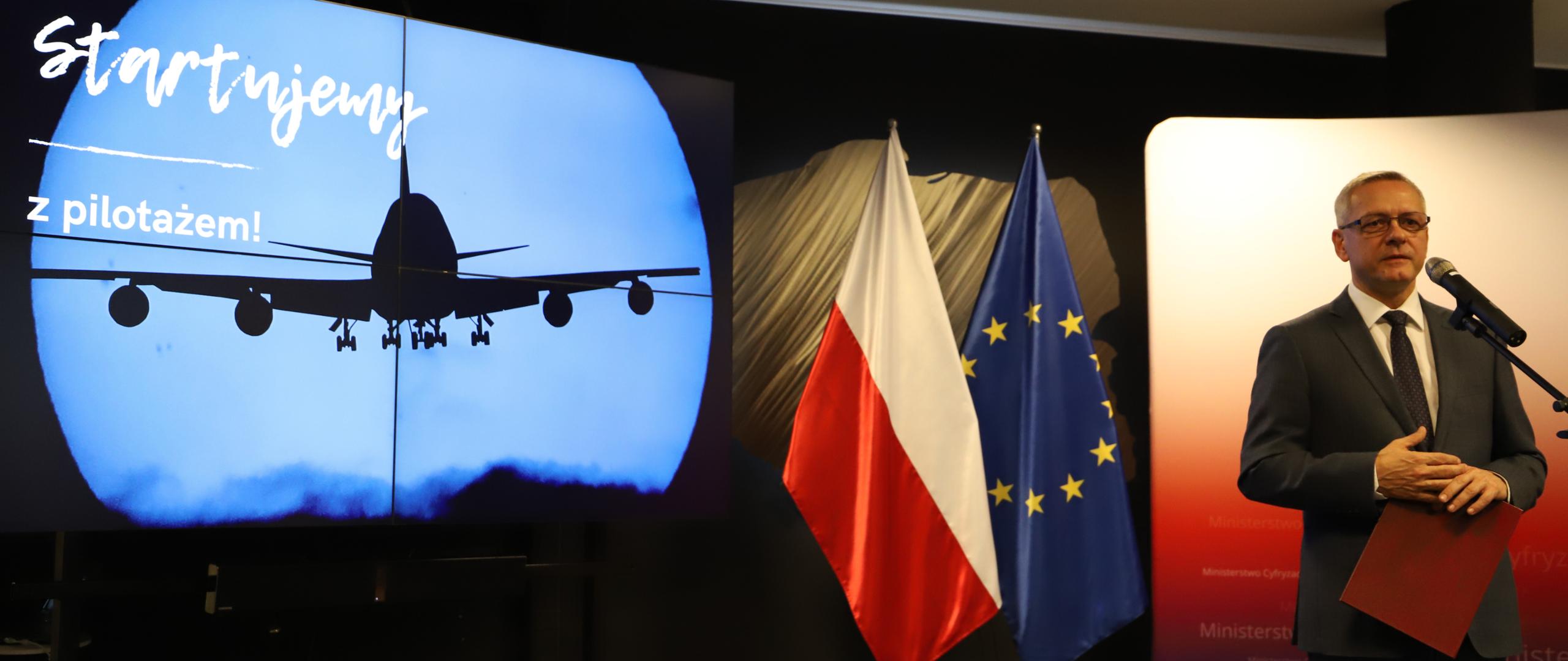 Minister Marek Zagórski podczas konferencji. W tle flagi: polska i unijna oraz ekran z prezentacją. Wyświetlany slajd pokazuje wznoszący się samolot, napisy: startujemy z pilotażem!