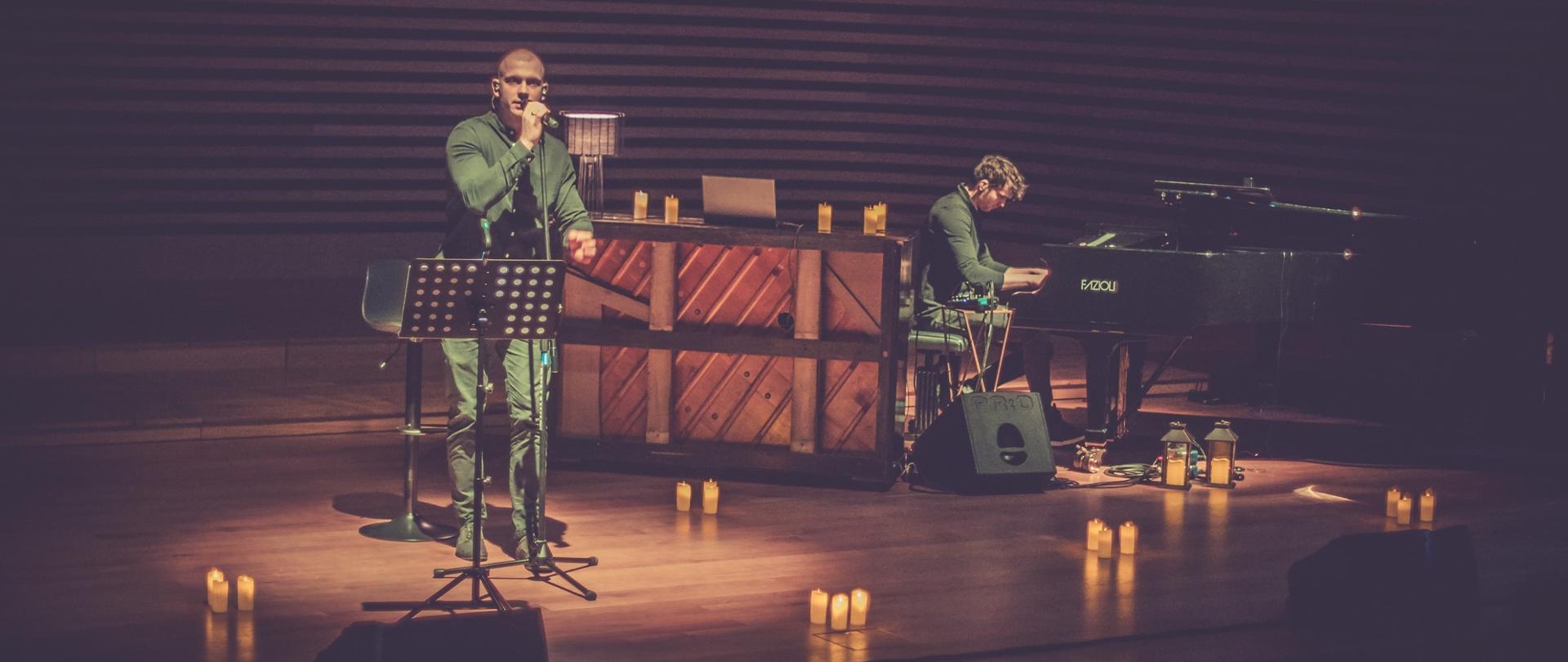 Mężczyzna stoi przy mikrofonie, po prawej mężczyzna gra na fortepianie, na scenie w różnych miejscach ustawione świece wprowadzające klimatyczny nastrój
