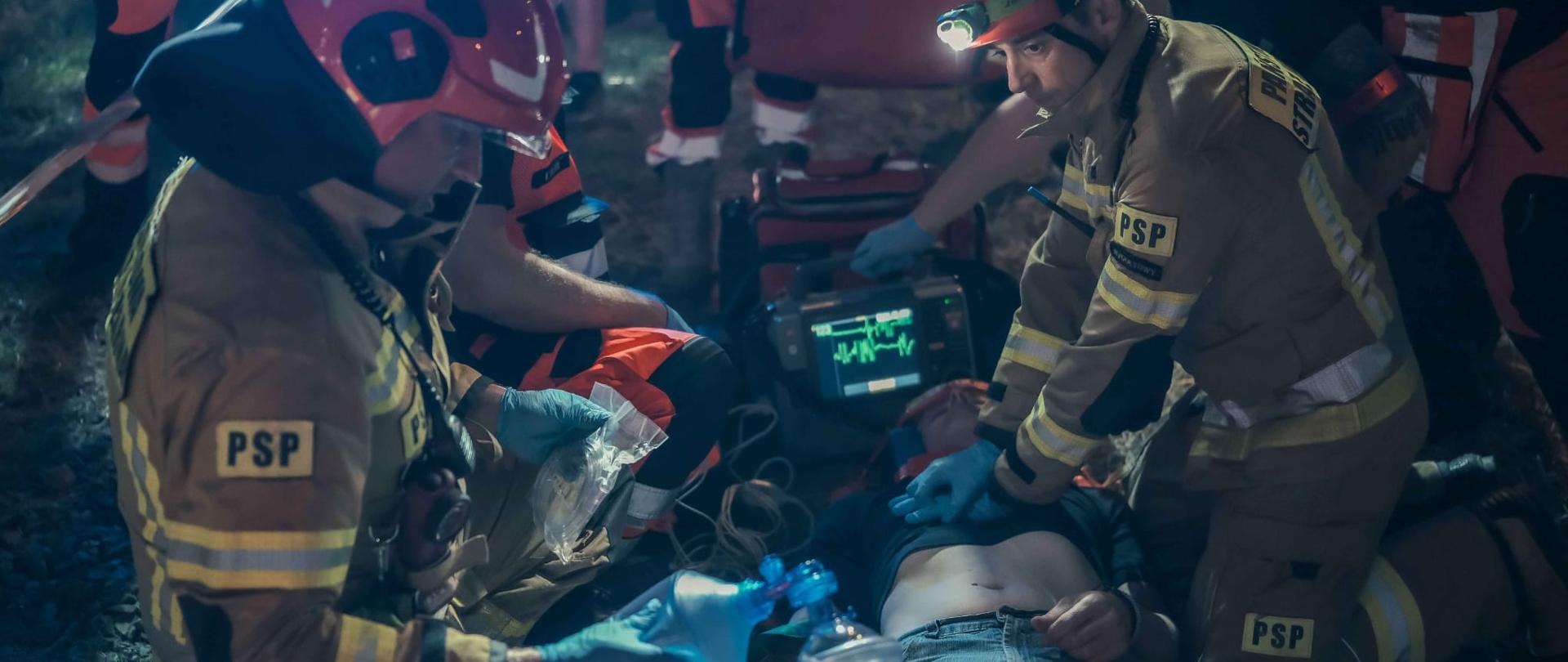 Fotografia przedstawia przykładowe działania PSP podczas wdrażania procedur i technik ratownictwa medycznego. Na fotografii widoczny leżący strażak pozorujący poszkodowanego, dookoła niego klęczą strażacy wykonując odpowiednie czynności ratownicze z użyciem specjalistycznego sprzętu.