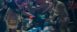 Fotografia przedstawia przykładowe działania PSP podczas wdrażania procedur i technik ratownictwa medycznego. Na fotografii widoczny leżący strażak pozorujący poszkodowanego, dookoła niego klęczą strażacy wykonując odpowiednie czynności ratownicze z użyciem specjalistycznego sprzętu.