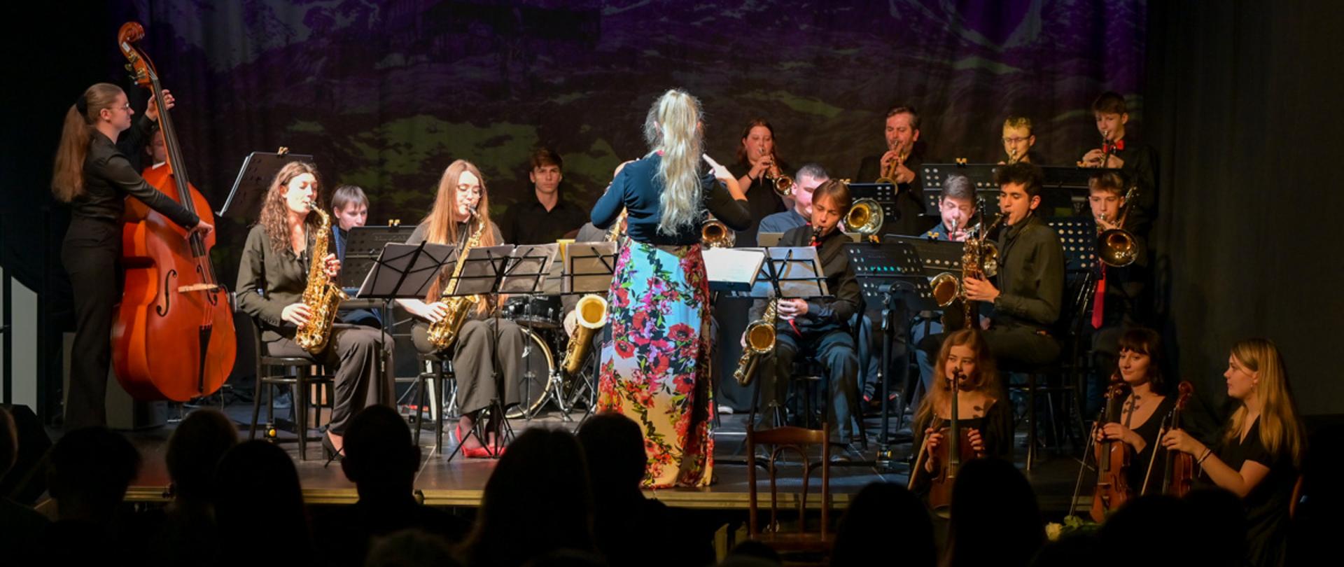 Zdjęcie na scenie Teatru Witkacy. Na pierwszym planie w mroku publiczność. Przed scena z prawej strony stoją trzy skrzypaczki. Na scenie big band dyrygowany, a przed nim dyrygentka. Z lewej strony stoi kontrabasistka i gra na kontrabasie. Big band złożony z uczniów i nauczycieli instrumentów dętych siedzi i gra na swoich instrumentach. Przed nimi pulpity, na których położone są nuty.