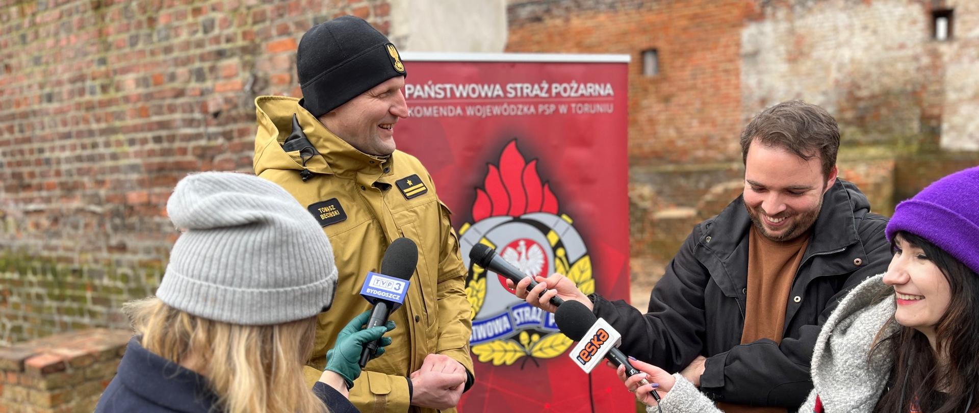 Strażak, a przed nim kilkoro dzienikarzy z mikrofonami. W tle baner firmowy, dalej mur średniowieczny Torunia.