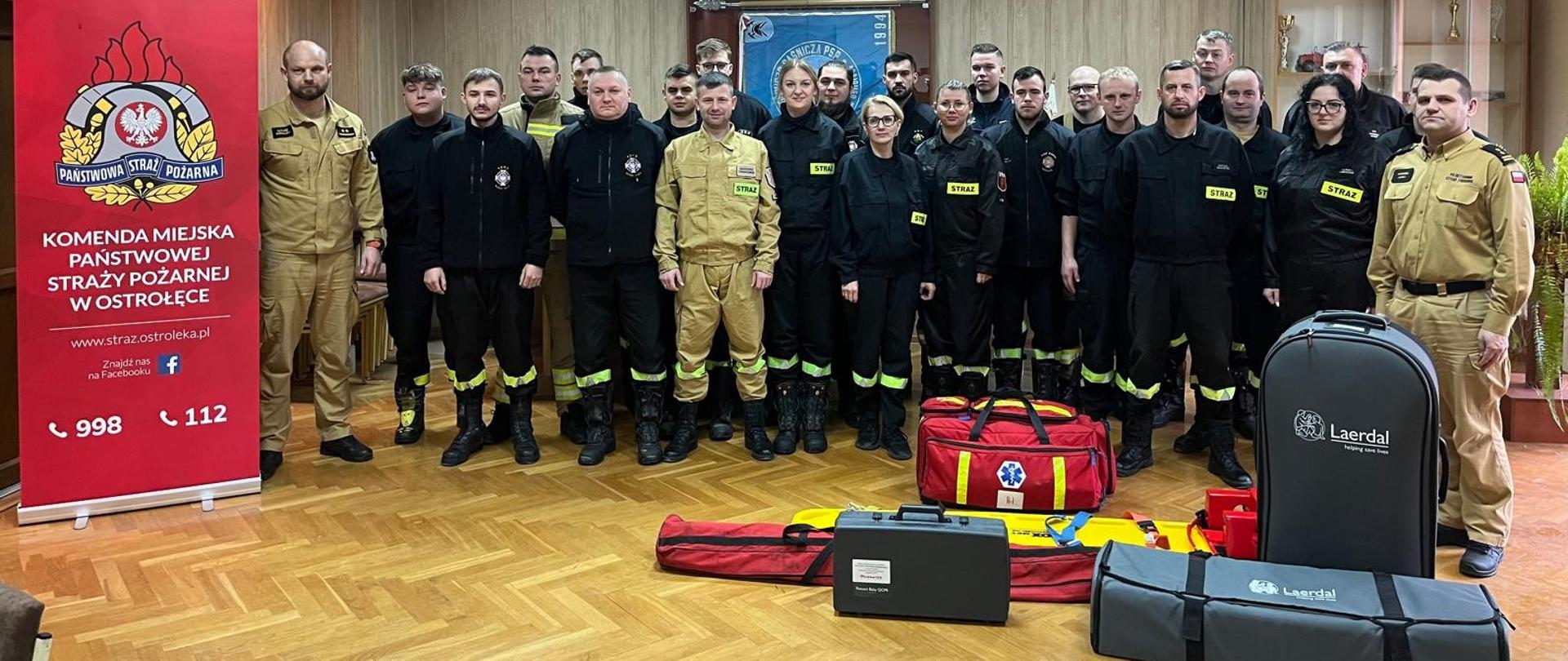 Na zdjęciu stoi duża grupa strażaków w umundurowaniu, ustawiona w kilku rzędach wewnątrz sali szkoleniowej. Po lewej stronie znajduje się czerwony roll-up z logo Państwowej Straży Pożarnej i nazwą Komendy Miejskiej PSP w Ostrołęce. Przed grupą, na podłodze, ułożony jest sprzęt szkoleniowy i medyczny, w tym torby ratownicze oraz pokrowce z logotypem Laerdal. W tle widać drewniane ściany, pamiątkową tablicę oraz przeszkloną gablotę z trofeami. Grupa strażaków na zdjęciu pamiątkowym po egzaminie z kursu kwalifikowanej pierwszej pomocy.