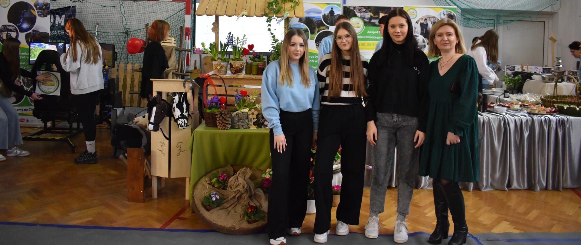 Trzy młode kobiety i jedna starsza pozują do zdjęcia na tle stoiska z kwiatami i drewnianymi dekoracjami w hali sportowej. Nad nimi wisi kosz do koszykówki.
