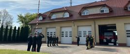 Na zdjęciu widać 14-stu strażaków. 10 z nich stoi w dwóch pięcioosobowych szeregach, zwróconych twarzami do siebie. Między nimi znajduje się strażak (dowódca) zwrócony twarzą do masztu z flagą znajdującym się na przeciw niego. Flagę polski na maszt wciąga 3 strażaków.