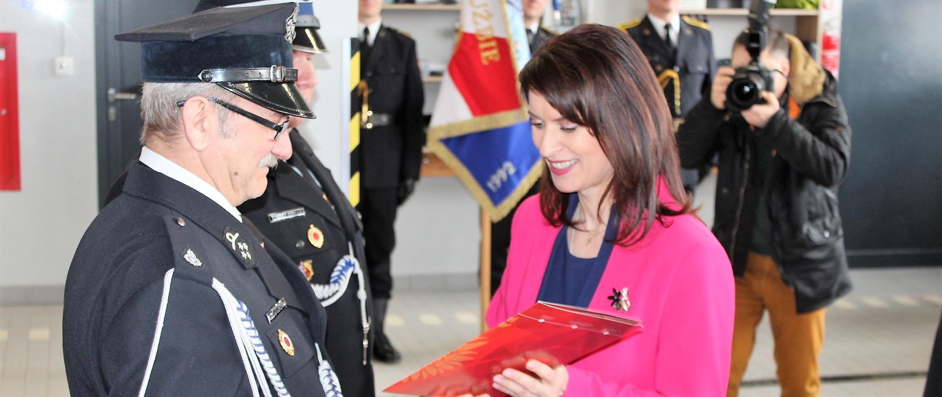 Pani Poseł w różowym kostiumie wręcza akt przekazania pojazdu druhom OSP. Strażacy w mundurach wyjściowych. W tle poczet sztandarowy i fotograf.