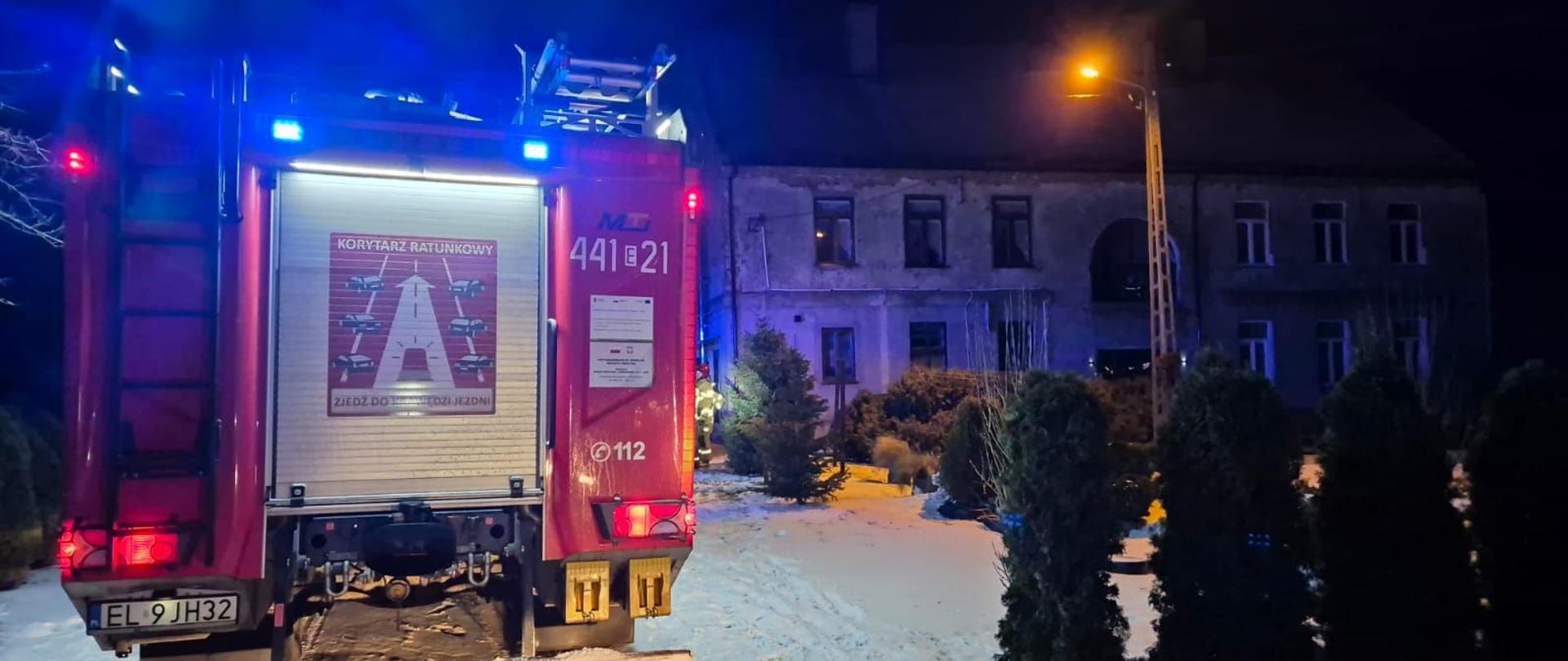 Na zdjęciu widać wóz straży pożarnej stojący nocą na zaśnieżonym terenie, z włączonymi niebieskimi światłami ostrzegawczymi. W tle widoczny jest murowany budynek z kilkoma oknami, częściowo oświetlony latarnią uliczną. Przed budynkiem rosną krzewy i małe drzewa, a ziemię pokrywa śnieg.
