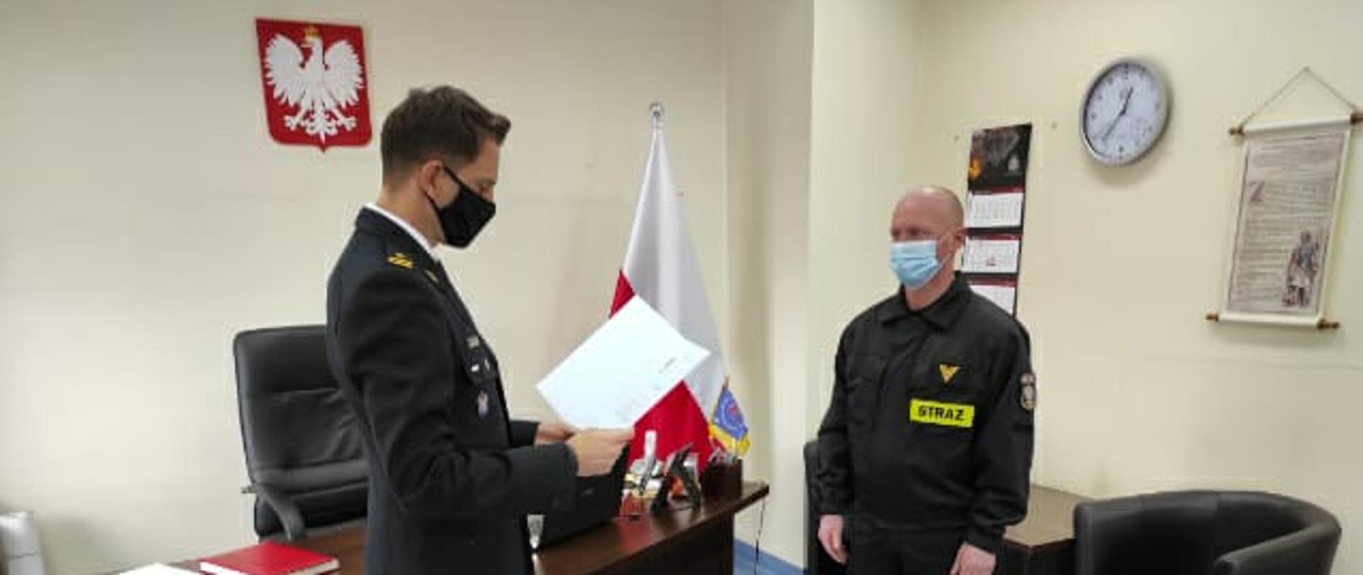 Na zdjęciu Komendant Powiatowy PSP w Myśliborzu wręczający strażakowi gratulacje. Obaj znajdują się w gabinecie. Na ścianie wisi godło Rzeczypospolitej Polskiej.