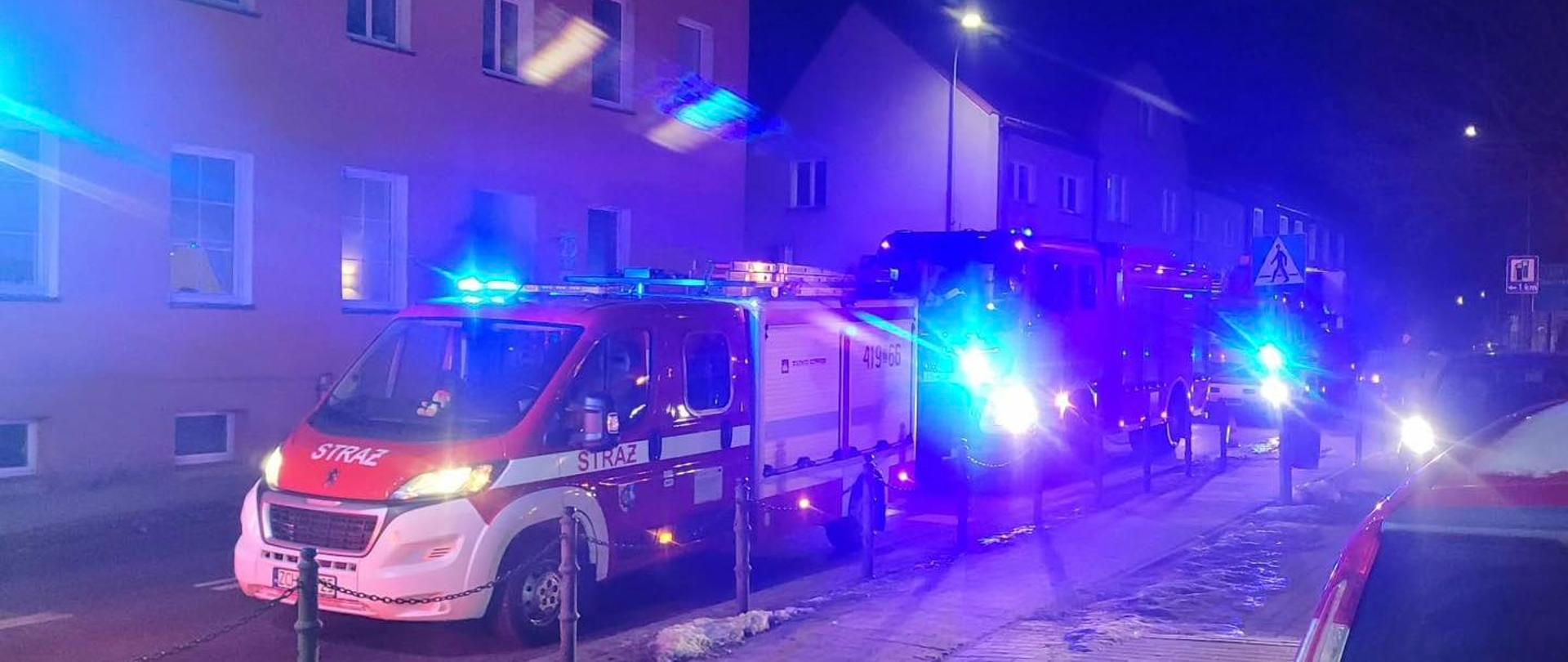 Trzy wozy straży pożarnej z włączonymi niebieskimi światłami stoją nocą na ulicy między budynkami mieszkalnymi; na chodniku widać śnieg i zaparkowane samochody.