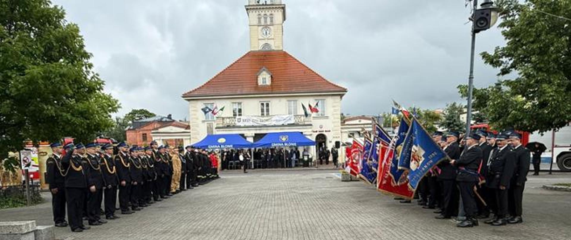 Widok ogólny na uroczystość. Po dwóch stronach placu stoją strażacy w mundurach wyjściowych. Strażacy oo prawej stronie zdjęcia prezentują sztandary. W centralnej części zdjęcia widoczny jest zabytkowy budynek, przed którym stoją niebieskie namioty. Pod zadaszeniem namiotów goście uroczystości. Ponad wszystkimi widoczne pochmurne burzowe niebo. 