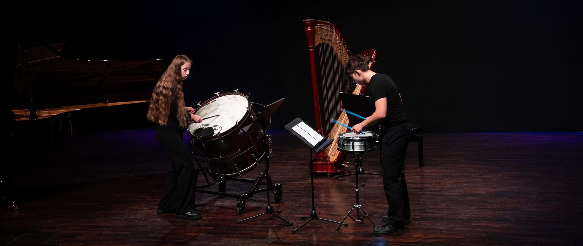 Zdjęcie przedstawia dwoje uczniów wykonujących wspólnie utwór na dwa instrumenty perkusyjne, w tle fortepian i harfa