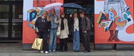Laureatki konkursu wraz z opiekunką pod parasolami pozują przed wejściem do budynku na tle kolorowych plakatów