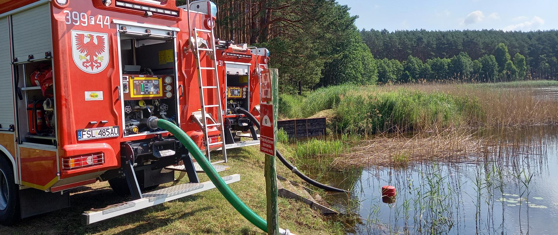Dwa samochody gaśnicze ustawione tyłem do zbiornika wodnego. Z autopomp pojazdów wyprowadzone do wody węże ssawne.