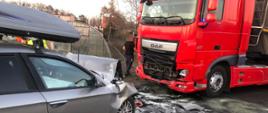 Czerwony samochód ciężarowy stoi z uszkodzonym przodem przed nim samochód osobowy srebrny z bagażnikiem na dachu z uszkodzonym przodem pomiędzy pojazdami plama posypana sorbentem 