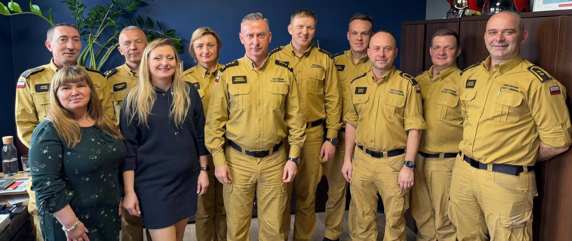 Na zdjęciu widoczna jest grupa funkcjonariuszy Państwowej Straży Pożarnej oraz pracowników cywilnych, zgromadzonych w pomieszczeniu biurowym. Strażacy ubrani są w jednolite, służbowe mundury koloru piaskowego, z oznaczeniami stopni i przynależności państwowej. W tle znajduje się godło Rzeczypospolitej Polskiej umieszczone na ścianie, a także elementy wyposażenia biura, w tym szafa oraz sprzęt ochronny (hełmy strażackie).