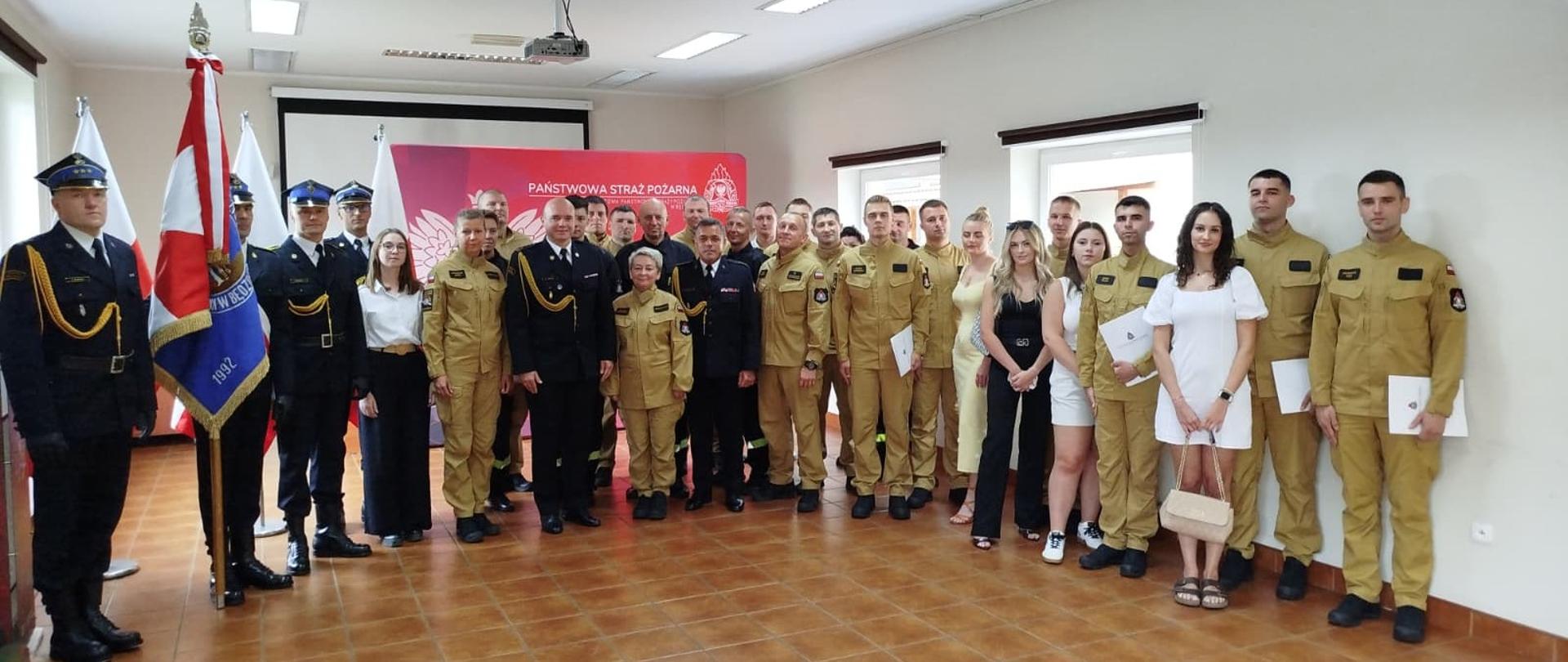 Na zdjęciu widać grupę osób pozujących do wspólnej fotografii w uroczystej sali. Na pierwszym planie i po bokach stoją strażacy w różnych mundurach – część w czarnych galowych z czapkami, inni w jasnożółtych bojowych. W środku znajdują się również osoby w cywilnych ubraniach, głównie kobiety w eleganckich kreacjach, prawdopodobnie zaproszeni goście lub rodziny strażaków.
W tle widnieje baner z napisem „Państwowa Straż Pożarna” i godłem orła, obok znajdują się także flagi Polski oraz sztandar strażacki. Wydarzenie wygląda na uroczystość wręczenia awansów, odznaczeń lub ślubowania strażaków.
Cała grupa prezentuje się uroczyście i oficjalnie, a atmosfera jest podniosła i pełna dumy.