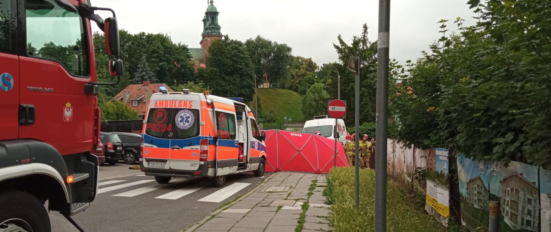 Na zdjęciu widać przód samochodu pożarniczego przed którym stoi Karetka Pogotowia. Przed karetką rozwinięty jest czerwony parawan strażacki osłaniający miejsce zdarzenia. W tle widać gnieźnieńską katedrę