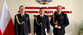 Trzech strażaków w mundurach galowych na tle napisu Wojewódzka Komenda Państwowej Straży Pożarnej w Szczecinie, nad napisem logo PSP, z boku flaga biało-czerwona