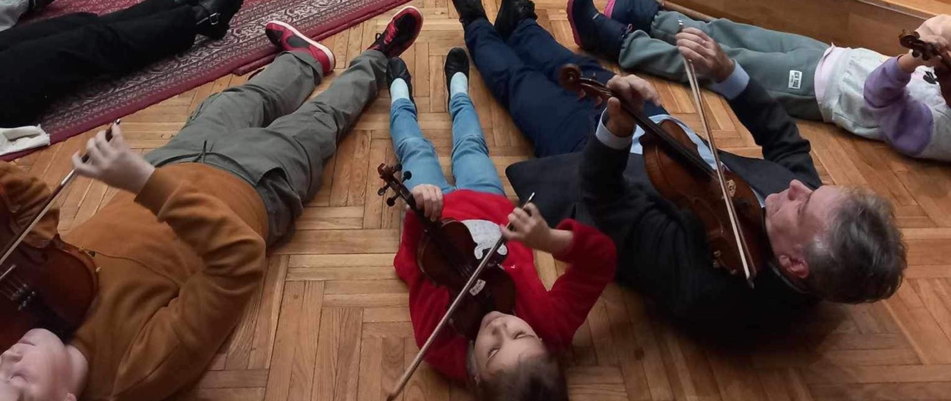 Uczestnicy warsztatów skrzypcowych wraz z prowadzącym podczas ćwiczenia w pozycji leżącej ze skrzypcami na parkiecie sali królewskiej PSM Mielec