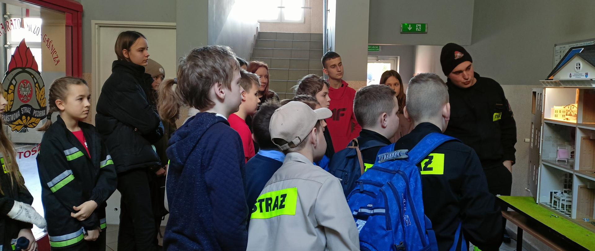 Zdjęcie przedstawia uczestników turnieju podczas lekcji edukacyjnej z wykorzystaniem symulatora zagrożeń pożarowych prezentowanego przez funkcjonariusza PSP w ubraniu koszarowym.