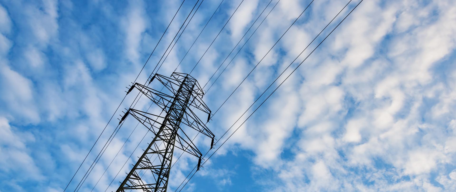 View of tall electricity pylons with multiple parallel wires extending overhead, photographed from a low angle against a bright sky.