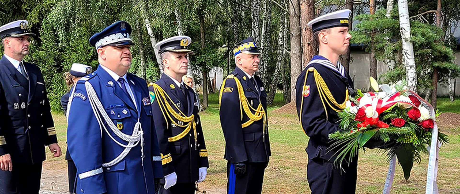 Żołnierz Marynarki Wojennej trzyma wiązankę za nim stoją przedstawiciele służb mundurowych Policji Straży Granicznej Państwowej Straży Pożarnej.